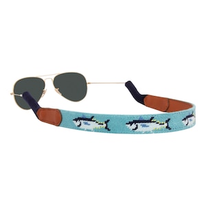 May include: A light blue sunglass strap with a brown leather accent and a repeating pattern of yellowfin tuna fish.