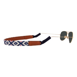May include: A blue and white patterned eyeglass strap with brown leather accents. The strap is attached to a pair of gold-rimmed sunglasses with dark lenses.