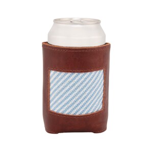 May include: Brown leather can cooler with a blue and white striped fabric patch. The cooler is designed to hold a standard-sized beverage can.