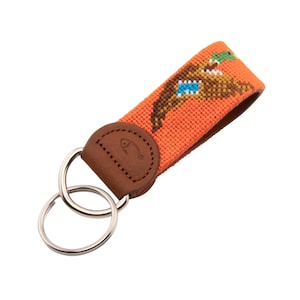 May include: Orange key chain with a brown leather strap and a double ring. The strap features an embroidered design of a duck with a blue head and green bill.