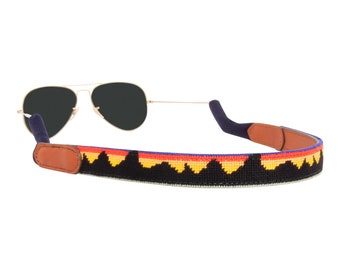 May include: A black and white patterned sunglass strap with a red, orange, and yellow mountain design. The strap is made of woven fabric and has brown leather ends.