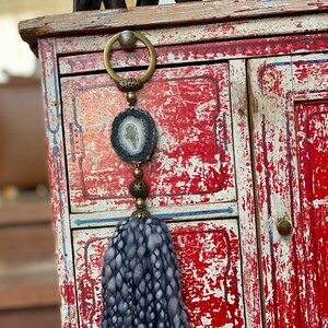 Könnte beinhalten: Ein grauer und weißer Achat-Steinanhänger mit einem Quasten, der an einem Messingring hängt. Die Quaste besteht aus grauem Garn und hängt an einem roten und weißen Schrank mit Abnutzungsspuren.