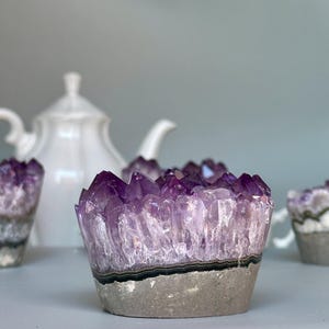 Puede incluir: Cuatro portavelas de cristal de amatista, dos están en foco, uno está parcialmente en foco y uno está fuera de foco. Los portavelas tienen forma de flor con una base natural, rugosa y gris. Una tetera blanca está en el fondo.