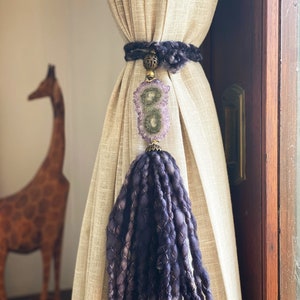 Może przedstawiać: Fioletowo-biały troczek do zasłon z frędzlami i dużym amuletem z kryształu ametystu. Troczek wykonany jest z grubego, skręconego sznurka i ma złoty metalowy pierścień.