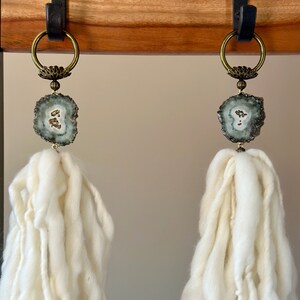 Puede incluir: Dos borlas decorativas con una estética natural y bohemia. Cada borla presenta un cuerpo grande de lana blanquecina, coronado con una rodaja de geoda y herrajes de latón. Las borlas cuelgan de una correa de cuero marrón oscuro y un anillo de latón.