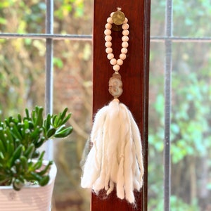 May include: A white tassel hanging from a wooden bead garland with a light brown agate pendant. The garland is attached to a gold-coloured metal ring.