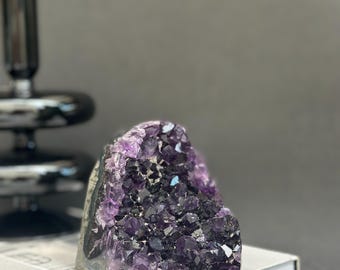 May include: A large, raw amethyst crystal geode with deep purple and black hues. The crystal is partially polished, revealing a sparkling interior. It sits on a closed book, with a black decorative object in the background.