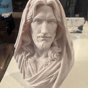 May include: A light beige, speckled bust sculpture depicting a figure with long, wavy hair and a beard, wearing a draped garment that covers the head and shoulders. The sculpture features detailed facial features and textured fabric folds.