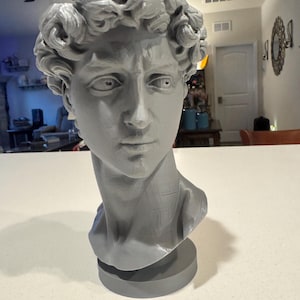 Könnte beinhalten: Graue Büste eines Mannes mit lockigem Haar und ernstem Gesichtsausdruck. Die Skulptur ist auf einem kreisförmigen Sockel montiert. Die Büste ist ein klassisches Kunstwerk.