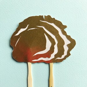 May include: Gold cupcake topper with a white swirl design and two wooden sticks.