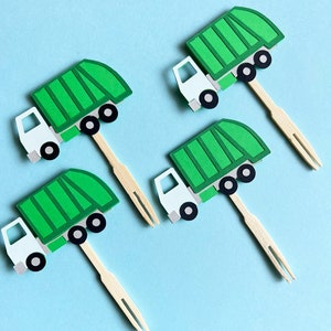 May include: Four green and white garbage truck cupcake toppers with wooden sticks. The trucks have a green body, white cab, and black wheels.