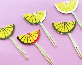 May include: Five yellow and red lemon slice cupcake toppers with gold foil accents. The lemon slices are on wooden skewers with two prongs at the end.