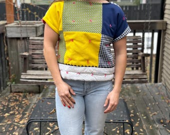 May include: A woman wearing a short-sleeved patchwork top with yellow, green, blue, and black fabric squares. The top has pink stitching and a white trim. She is wearing light blue jeans with a distressed knee.