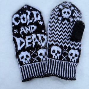 Op de afbeelding: Een paar zwart-witte gebreide wanten. Eén want toont de woorden "COLD AND DEAD" met schedelontwerpen. De andere want heeft een schedel- en kruisbottenpatroon. De wanten liggen op een wit oppervlak.