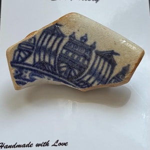 Può includere: Un frammento liscio e levigato di ceramica bianca con un disegno blu stampato a trasferimento raffigurante un veliero. Il testo Northumbrian Sea Pottery e Handmade with Love DoubleRByLynn è stampato sul supporto di cartone bianco retrostante.