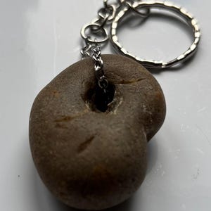 Può includere: Un portachiavi in pietra naturale con una catena e un portachiavi color argento. La pietra è di colore marrone arrotondato con un foro al centro. Il portachiavi è un accessorio unico e naturale.
