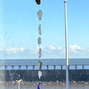 May include: A sea glass wind chime, crafted from pieces of white, green, and blue glass, hangs from driftwood. The chime is suspended against a backdrop of a blue sky and water.