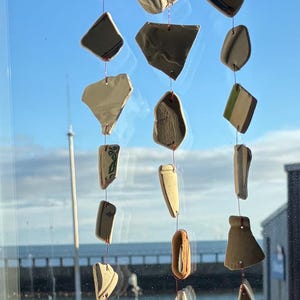 Puede incluir: Un móvil eólico artesanal hecho con piezas de cerámica recicladas. El móvil presenta varias formas y colores de cerámica, suspendidas de una rama natural con hilo rojo. El fondo muestra un cielo azul claro y una masa de agua.