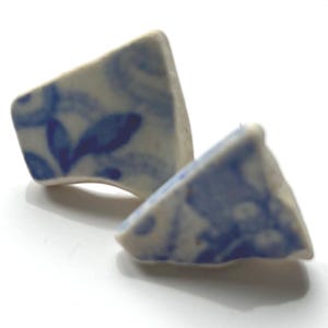 May include: Two ceramic earrings with a triangular shape. The earrings are white with a blue floral pattern. The earrings are displayed on a white surface.