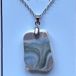 May include: A silver chain necklace with a rectangular sea pottery pendant. The pendant displays blue, grey, and white hues. The words "Northumbrian Sea Pottery" are printed above the necklace. The words "Handmade with Love" are also visible.