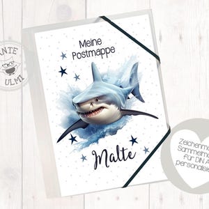 May include: A white folder with a blue shark illustration and the text "Meine Postmappe Malte" on the front. The folder is decorated with blue stars and a watercolour effect.