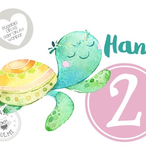 Könnte beinhalten: Eine Aquarell-Illustration einer grünen Meeresschildkröte mit einem gelben und orangefarbenen Panzer. Die Schildkröte trägt eine Blumenkrone und hat die Augen geschlossen. Der Text "Hani 2" ist in Grün und Rosa auf weißem Hintergrund geschrieben.