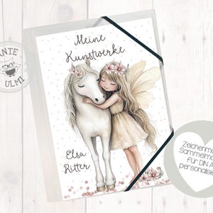 Könnte beinhalten: Ein weißes Einhorn mit einer rosa Blumenkrone steht neben einer Fee mit einer rosa Blumenkrone und einem weißen Kleid. Der Text "Meine Kunstwerke" steht über dem Einhorn und der Fee. Der Text "Elsa Ritter" steht unter dem Einhorn und der Fee. Der Text "Zeichenmappe Sammelmappe für DIN A4 personalisiert" steht auf einem weißen Kreis.