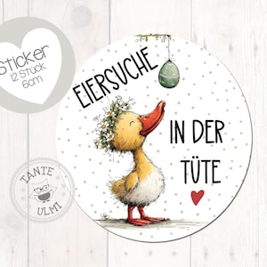 Könnte beinhalten: Ein runder Aufkleber mit einer Cartoon-Ente mit Blumenkranz, die zu einem hängenden grünen Ei aufblickt. Der Aufkleber trägt den Text "Eiersuche in der Tüte" und "Sticker 12 Stück 6cm". Ein Herz und das Tante Ulmi-Logo sind ebenfalls zu sehen.