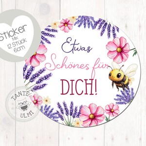 Peut inclure: Autocollant rond avec une bordure florale de fleurs roses et de lavande violette. L'autocollant porte le texte "Etwas Schönes für DICH !" et une abeille de dessin animé. Un petit autocollant en forme de cœur est également présent.