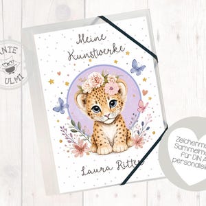 Könnte beinhalten: Eine weiße A4-Sammelmappe mit schwarzem Gummizug, verziert mit einer Illustration eines Leopardenbabys mit Blumenkranz, Schmetterlingen und Sternen sowie dem aufgedruckten Text Meine Kunstwerke und Laura Ritter auf der Vorderseite.