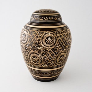 May include: Black and gold metal urn with an intricate floral design. The urn has a lid and a round body.