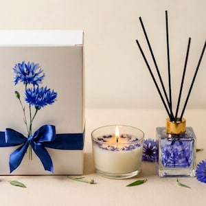 Puede incluir: Una caja de regalo con una cinta azul y un diseño floral, una vela encendida y un difusor de varillas con varillas negras. La vela y el difusor contienen pétalos de flores azules. La escena está decorada con flores azules y hojas verdes.