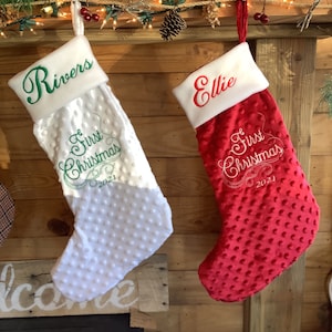 May include: Two personalized Christmas stockings, one white with green embroidery and the name "Rivers" and the other red with white embroidery and the name "Ellie". Both stockings have the text "1st Christmas 2021" embroidered on them.
