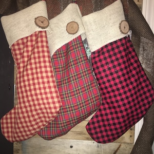 May include: Three red and white Christmas stockings with burlap cuffs and wooden button accents. The stockings are hanging on a rustic wooden background.