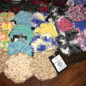 Op de afbeelding: Een verzameling kleurrijke gehaakte schuursponsjes, sommige met een pluche textuur en andere met een gladde textuur. De schuursponsjes zijn in een cirkel op een houten oppervlak gerangschikt. Een klein label met de tekst "Made 2 Motown Creations" is zichtbaar in de rechterbenedenhoek van de afbeelding.