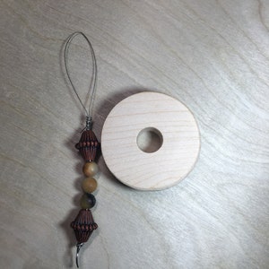 May include: A handmade wooden yarn swift with a decorative beaded needle threader. The threader features brown, tan, and black beads, and a wire loop. The yarn swift is a light wood color with a hole in the center.