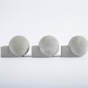 May include: Three grey concrete sphere sculptures, each with a spherical top and a square base. The spheres are set on a white background. The sculptures have a modern, minimalist design.