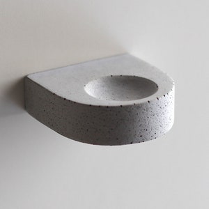 May include: A small, white concrete wall-mounted shelf with a rounded, indented area in the center. The shelf has a speckled texture.