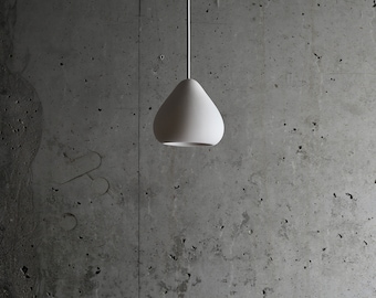 May include: A white teardrop-shaped pendant light fixture hangs from a ceiling against a concrete wall.