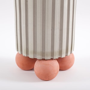 May include: A white concrete planter with a fluted design sits on four terracotta spheres. The planter is a cylindrical shape and the spheres are a slightly darker shade of orange.