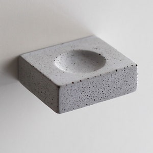 Puede incluir: Un pequeño plato rectangular de hormigón con una hendidura redonda en el centro. El plato tiene un acabado gris claro moteado.