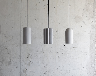 May include: Three contemporary pendant lights in grey and white hang against a concrete wall. The lights have cylindrical and rounded forms, with black cords. The central light has a ribbed design. The lights are suspended.