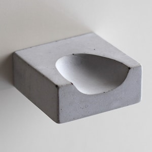 Puede incluir: Una pequeña repisa de pared de hormigón blanco con un área curva y hundida para guardar objetos pequeños.