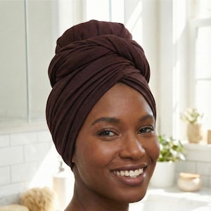 May include: A woman wearing a dark brown hair wrap. The wrap is made of a soft, draped fabric and is styled in a turban-like fashion. The woman is smiling, and the background includes a bathroom setting with a sink and toiletries.