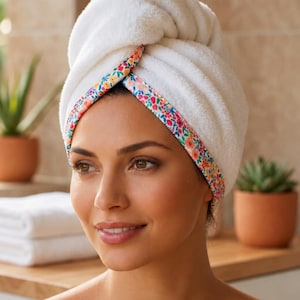 Smiling woman wearing a white cotton hair towel wrap with a colorful floral trim. Soft and absorbent hair drying turban for post-shower and spa routines.