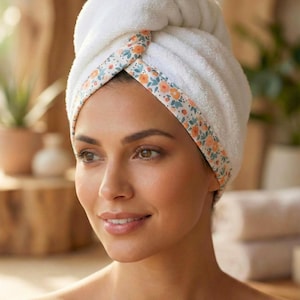 Puede incluir: Una toalla para el cabello de color blanco con un borde floral de flores naranjas y azules. La tela tiene una textura suave y afelpada, envuelta firmemente alrededor de la cabeza. El texto "Style - 12" aparece impreso en negro abajo.
