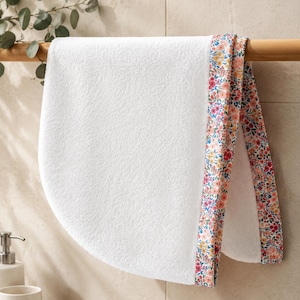 White terry cloth hair towel with a floral patterned trim. The trim features pink, yellow, and purple flowers with green leaves on a white background. The towel is hanging on a wooden rack.