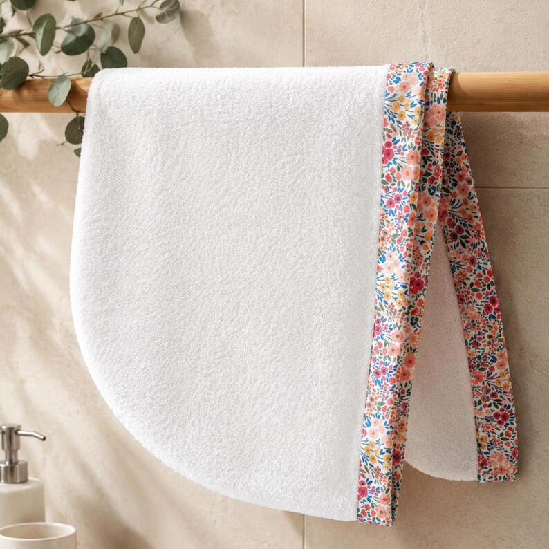 White terry cloth hair towel with a floral patterned trim. The trim features pink, yellow, and purple flowers with green leaves on a white background. The towel is hanging on a wooden rack.