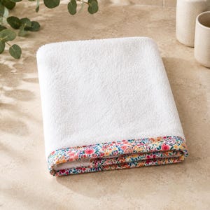 Folded white towel with a floral patterned border. The border features a colorful design with pink, yellow, and blue flowers on a white background. The towel is made of a soft, absorbent material.