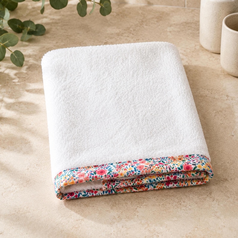 Folded white towel with a floral patterned border. The border features a colorful design with pink, yellow, and blue flowers on a white background. The towel is made of a soft, absorbent material.